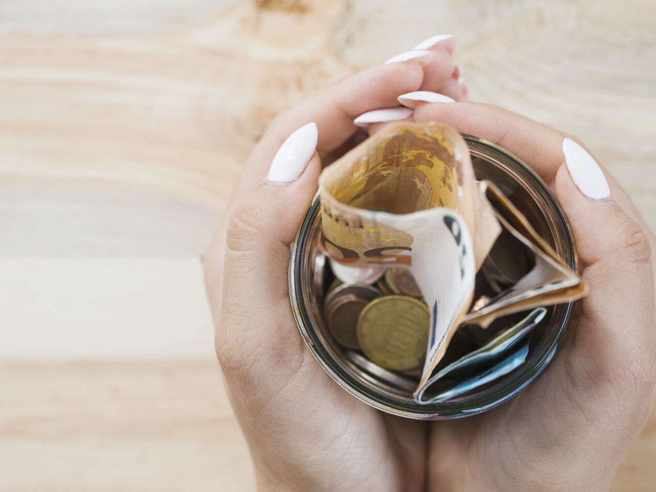 main-femme-tenant-bocal-verre-billets-pieces-euros-fond-bois